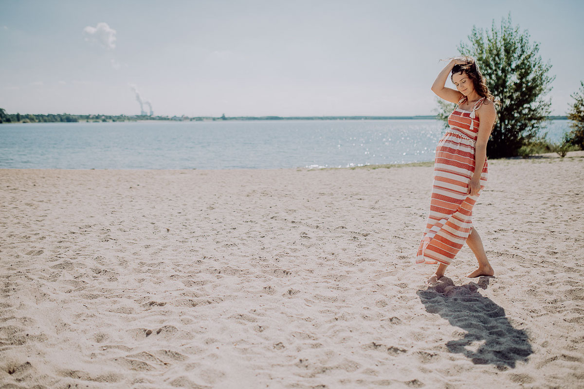 Babybauch Shooting, Foto einer schwangeren Frau am Strand mit Wind, Sonne und blauem Himmel