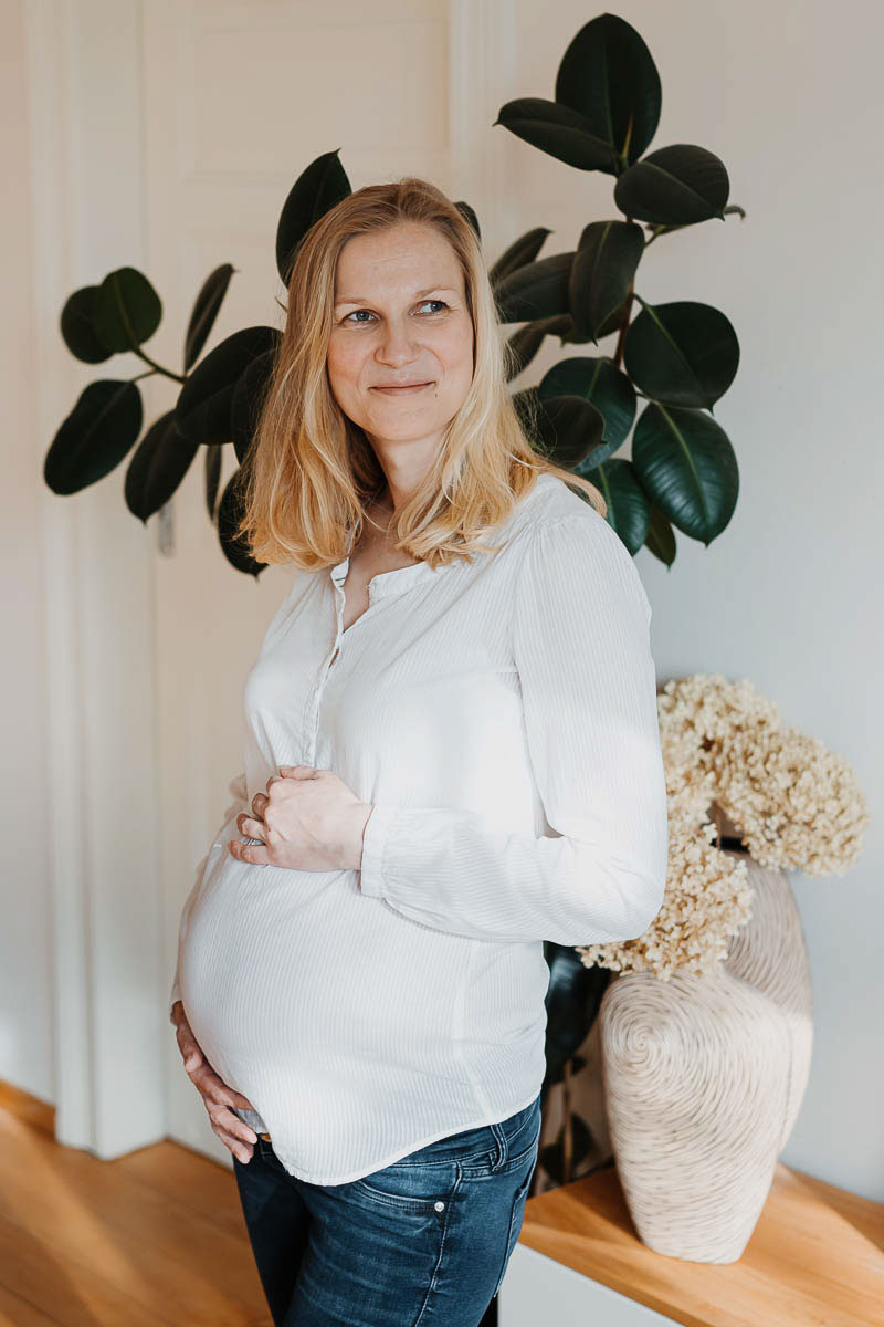 Babybauch Shooting schwangere Frau in weißem Oberteil schaut zum Fenster